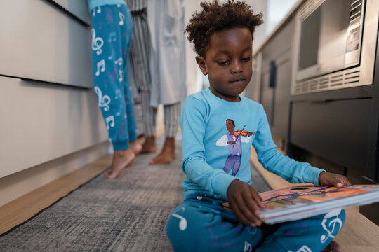 Cute boy in pajamas reading book on kitchen floor
