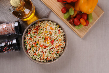 Couscous with vegetables in a rustic bowl surrounded by tomatoes, peppers and a glass of olive oil....