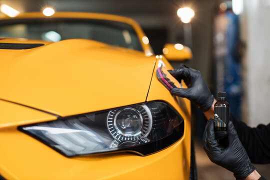 Cropped close up image of male hand in protective black glove, holding sponge with solid carnauba wax, and polishing hood of luxury yellow car at professional detailing workshop. Car detailing.