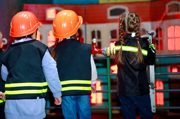 Two little girls dressed as firefighters play role-playing games and water a toy house with a hose, girls in a hard hat, children's games.
