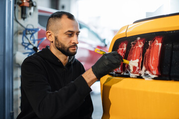 Close up photo of young male worker in black protective gloves cleaning tail light in foam with special brush. Sport yellow luxury car wash and detailing. Washing machine at the station.