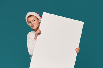 Surprised teen boy  in a Santa Claus hat and party glasses on a turquoise background. holds a white...