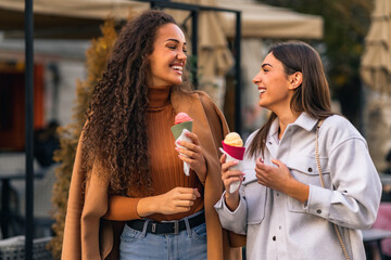 Two girls are strolling, talking and eating cone ice cream, and having much fun in the town.