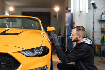 Auto detailing service, polishing of the car. Side view of young man worker in black clothes, polishing yellow car body with orbital polisher and wax to eliminate contaminants from the surface.
