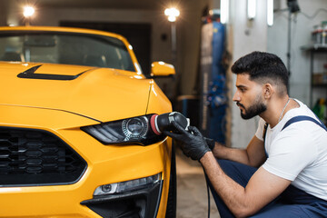 Young professional bearded male worker in overalls and white t-shirt with orbital polisher in auto...