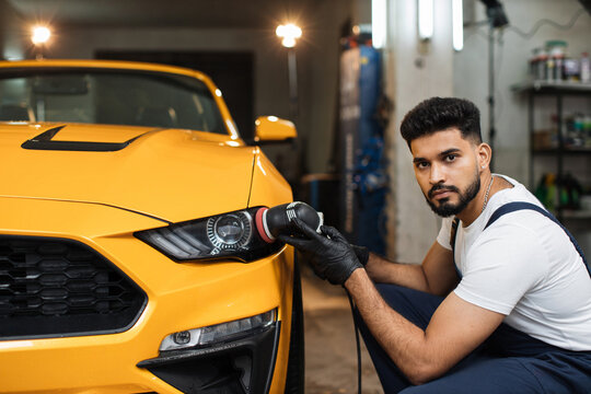 Young professional bearded male worker in overalls and white t-shirt with orbital polisher in auto service polishing headlights of yellow luxury car. Detailing and polishing of car headlights.