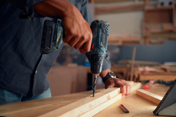 Close-up of young man using drill on wooden plank on top of table in woodworking factory