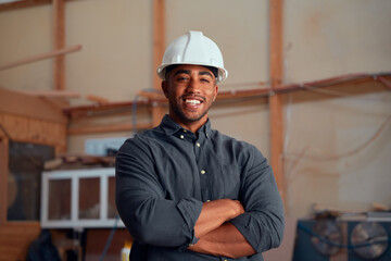 Portrait of happy young man in safety helmet looking at camera with arms crossed at woodworking...