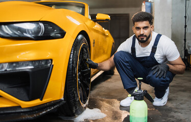 Handsome young bearded male worker in uniform, cleaning the wheel rim of modern yellow sport car by...