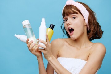 a shocked, surprised woman stands on a blue background, wrapped in a white towel and holding a set of jars with facial care cosmetics in her hands, her mouth wide open with emotions