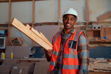 Young man wearing protective workwear smiling while carrying wood at woodworking factory