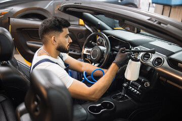 Handsome male cleaner in overalls, white t-shirt and protective gloves, removing dirt from car...