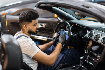 Handsome bearded young man in uniform and protective gloves, cleaning car interior and steering wheel using microfiber cloth, smiling at camera. Car detailing and valeting concept.