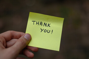 Close up on a yellow sticky note saying thank you  held by a hand. Dark background, bokeh