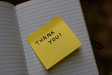 Close up on a yellow sticky note saying thank you glued on the page of an opened brown notebook.
