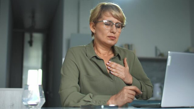 Adult blond woman in eyeglasses working laptop, feeling pain touching heart, risk of heartattack, nervous tension because of hard work in applications on pc, lack of knowledge but high responsibility