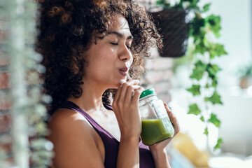 Happy woman drinking a healthy green smoothie standing in the living room at home.