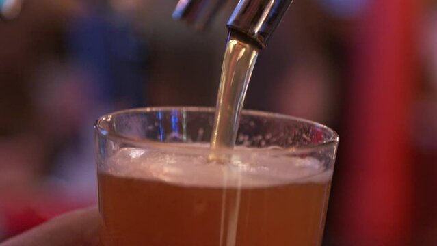 draft beer being poured into a clear glass using a metal draft keg in a dark and busy bar 