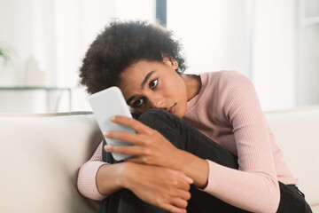 Sad depressed young african american woman with curly hair hugging her knees and looking at smartphone