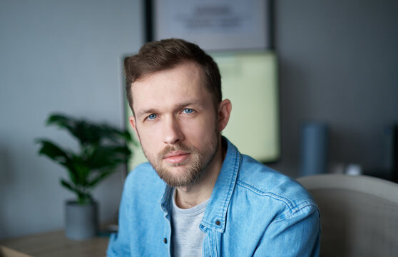 Young handsome man in denim shirt, positive emotion with computer monitor on background. Cheerful bearded male sitting in home office looking at camera with serious emotion. High quality image