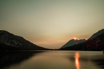 Toxaway Lake, ein alpiner See in Idahos Sawtooth Wilderness, gesehen an einem Sommertag im Morgengrauen, wenn die Sonne über den Bergen aufgeht und orangefarbenes Licht in der Wasseroberfläche reflektiert.