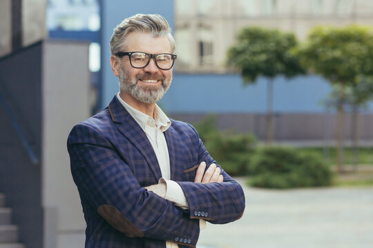 Portrait of mature gray-haired investor, senior man in glasses looking at camera and gesturing, businessman with crossed arms outside office building in business suit, successful boss banker.
