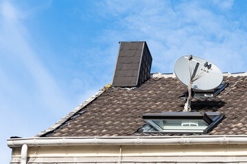A fragment of a tiled roof with satellite dish and a chimney against a blue sky with light clouds.