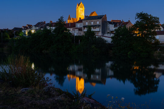Evening view of Dormans townscape with tall lighted bell tower of Saint-Hippolyte church rising above residential areas and reflected in water of Marne river in summer, France