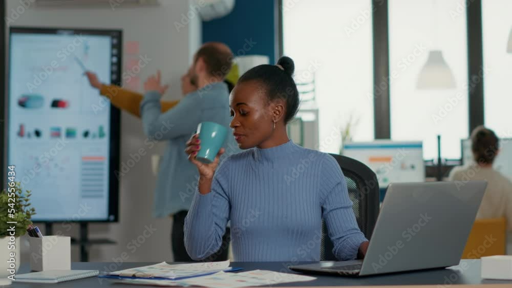 Poster African american woman taking a break from work using laptop to drink a sip of coffee or tea from cup in busy office. Casual startup employee working relaxed with statistics in business workplace.