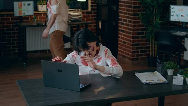 Brain dead spooky zombie working on modern computer. Brain-eating creppy dead walking corpse with bloody and deep wounds sitting at desk in office while trying to use laptop for reading documents.