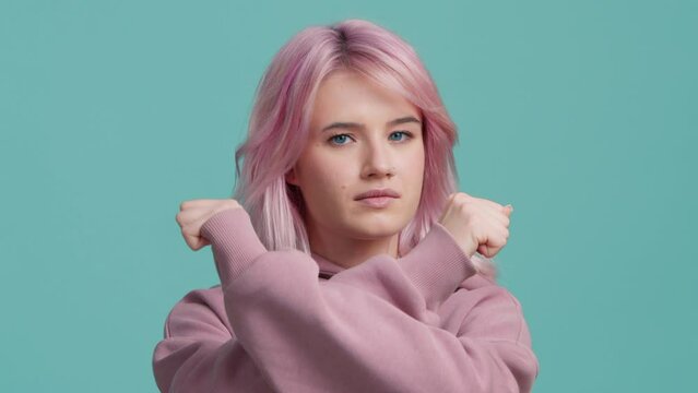 Say no holding folded crossed hands stop gesture. Slow motion displeased young student hipster girl with pink hair wearing casual hoodie isolated on pastel blue background 4K. People lifestyle concept