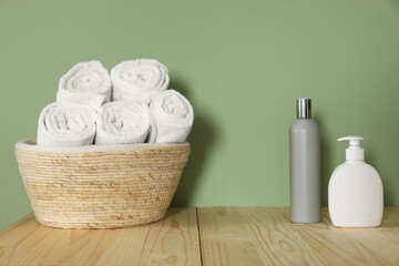 Fototapeta na wymiar Wicker basket with clean towels and cosmetic products on wooden countertop near green wall
