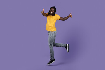 Fototapeta na wymiar Joyful black guy jumping on purple, showing thumb ups