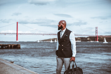 Fototapeta premium An outdoor shot with a fashionable handsome mature bald black man entrepreneur with a beautiful beard, bag in his hand, eyeglasses, and a suit with a vest; a red rope bridge in a defocused background