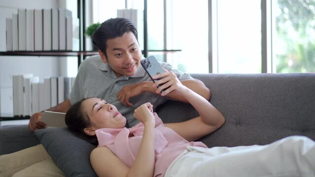 Happy cheerful Asian couple living together in living room in a free time in weekend, Asian girl laying down on sofa with her boyfriend and watching a video on smartphone.