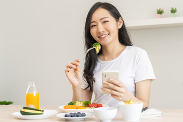 Dieting, asian young woman eating, holding fork at broccoli, diet plan nutrition with fresh vegetables salad, enjoy meal while using smartphone. Nutritionist of healthy, nutrition of weight loss.
