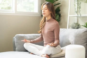 Fotobehang Air humidifier, calm asian young woman sitting on couch lotus pose put hands practice meditation do yoga exercise at home. Aromatherapy steam scent from essential oil diffuser in living room at home. © KMPZZZ
