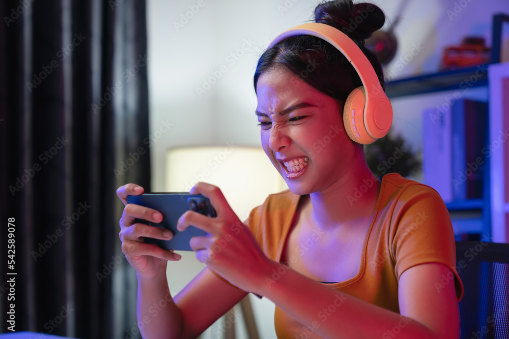 Wall mural excited young asian woman wearing headset and playing online game on smartphone at night modern hous