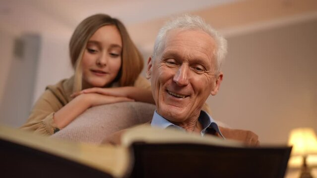Portrait of Caucasian positive senior man talking recalling memories watching old photo album with blurred teenage girl listening stories at background. Happy grandfather and granddaughter at home
