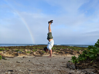 Man does Handstand on coral rocks on the beach with rainbow