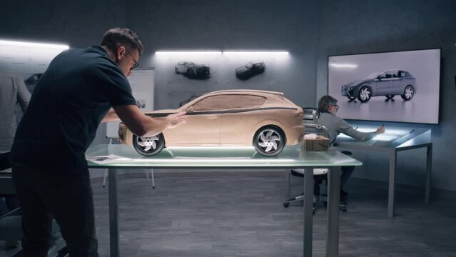 Two male automotive designers working on model of car in modern car design studio. One sculpting with plasticine clay and with rake, other working at the touch table. Future design of electric car.
