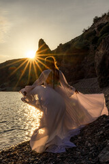 A mysterious female silhouette with long braids stands on the sea beach with mountain views, Sunset...
