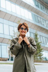 Smiling happy young asian model with short haircut wearing round stylish glasses and touching green shirt standing against modern architecture background and looking at camera outdoors