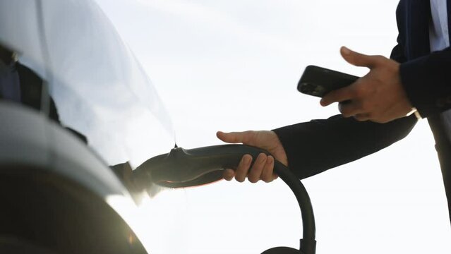 Unrecognizable businessman plugging electric car from charging station using app on smartphone. Man is plugging in power cord to an electric vehicle at sunset. Charging electric car
