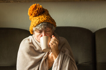 Elderly woman in ginger cap wrapped in blanket and drinking cup of tea. Old grandma feeling cold at...