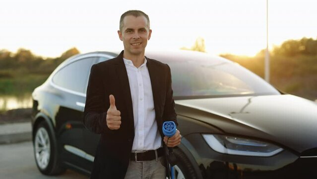 Portrait of caucasian businessman standing near electric charging station looking at camera and showing thumb up. Male holds a charging cable type 2 mennekes in his hands. Eco-friendly transport