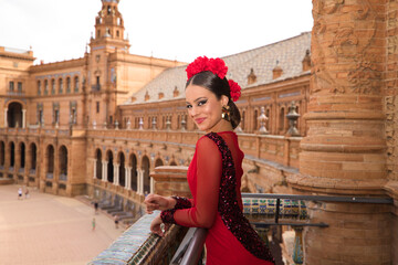 Beautiful teenage woman dancing flamenco on the balcony of a square in Seville. She wears a red dress with ruffles with a lot of art. Flamenco cultural heritage of humanity. - obrazy, fototapety, plakaty