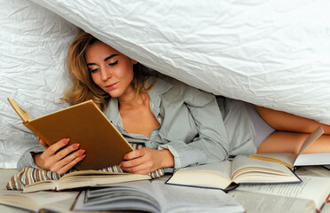 Cute blonde woman in white shirt on the bed in home bedroom, reads a book. Model under a blanket cover with a book. Many books in the foreground