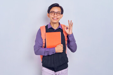 Happy smiling young Asian students with backpacks carrying books to school, showing okay sign isolated on gray background. Education in high school university college concept