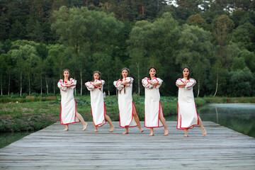 Young people in Slavic clothes  dancing on the pier near the lake. Kupala summer. - obrazy, fototapety, plakaty